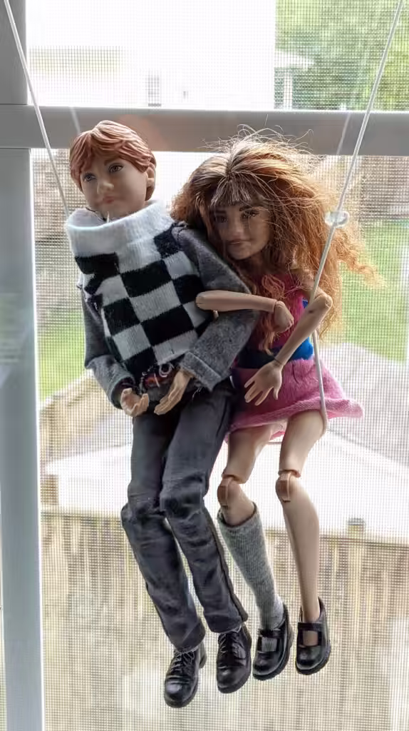 A photo of two dolls hanging on a string in front of a window as if on a swing